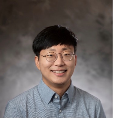 Headshot of Changwoo Lee, smiling, in round glasses and a blue button-down shirt against a mottled grey studio background.