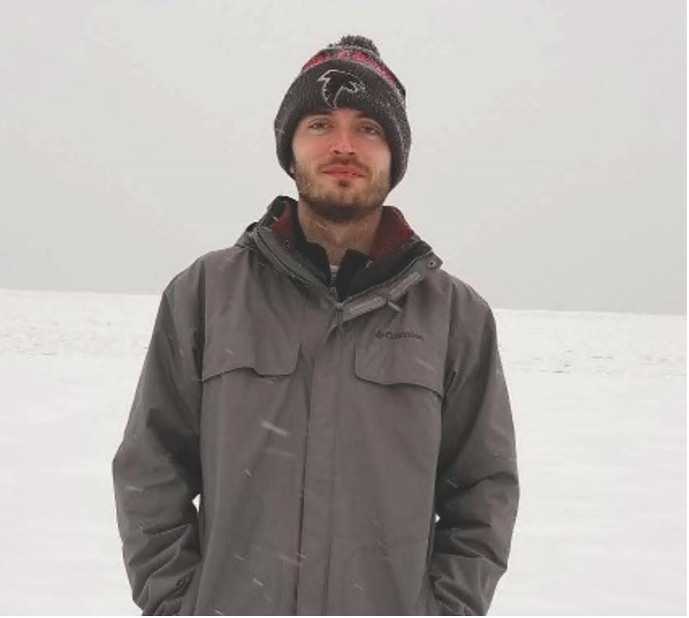Jacob Martin in a knit winter hat and grey jacket, standing in falling snow on an open snowy field.